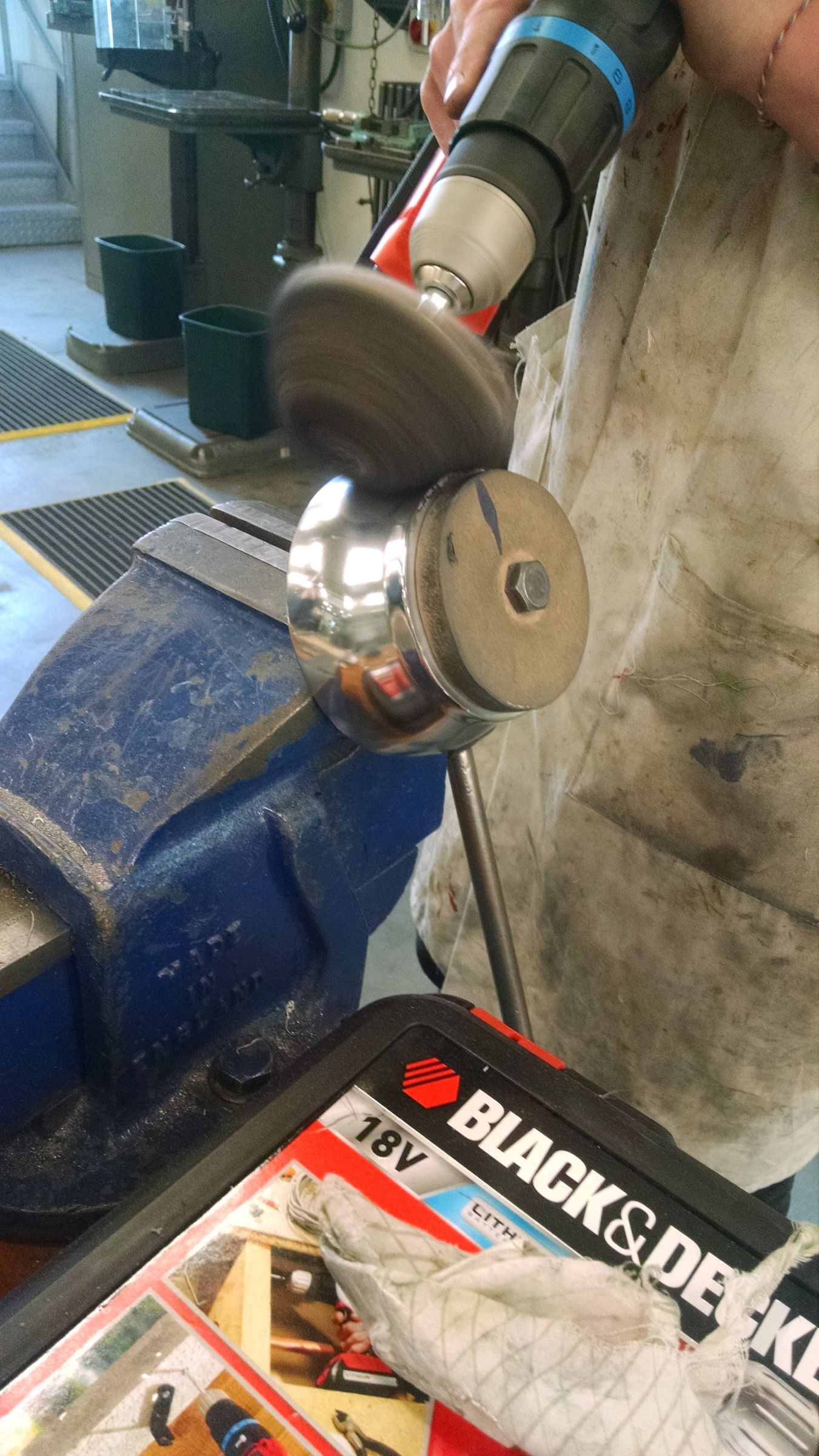 A cylindrical metal part being finished on a bench grinder, with a magazine visible in the background.