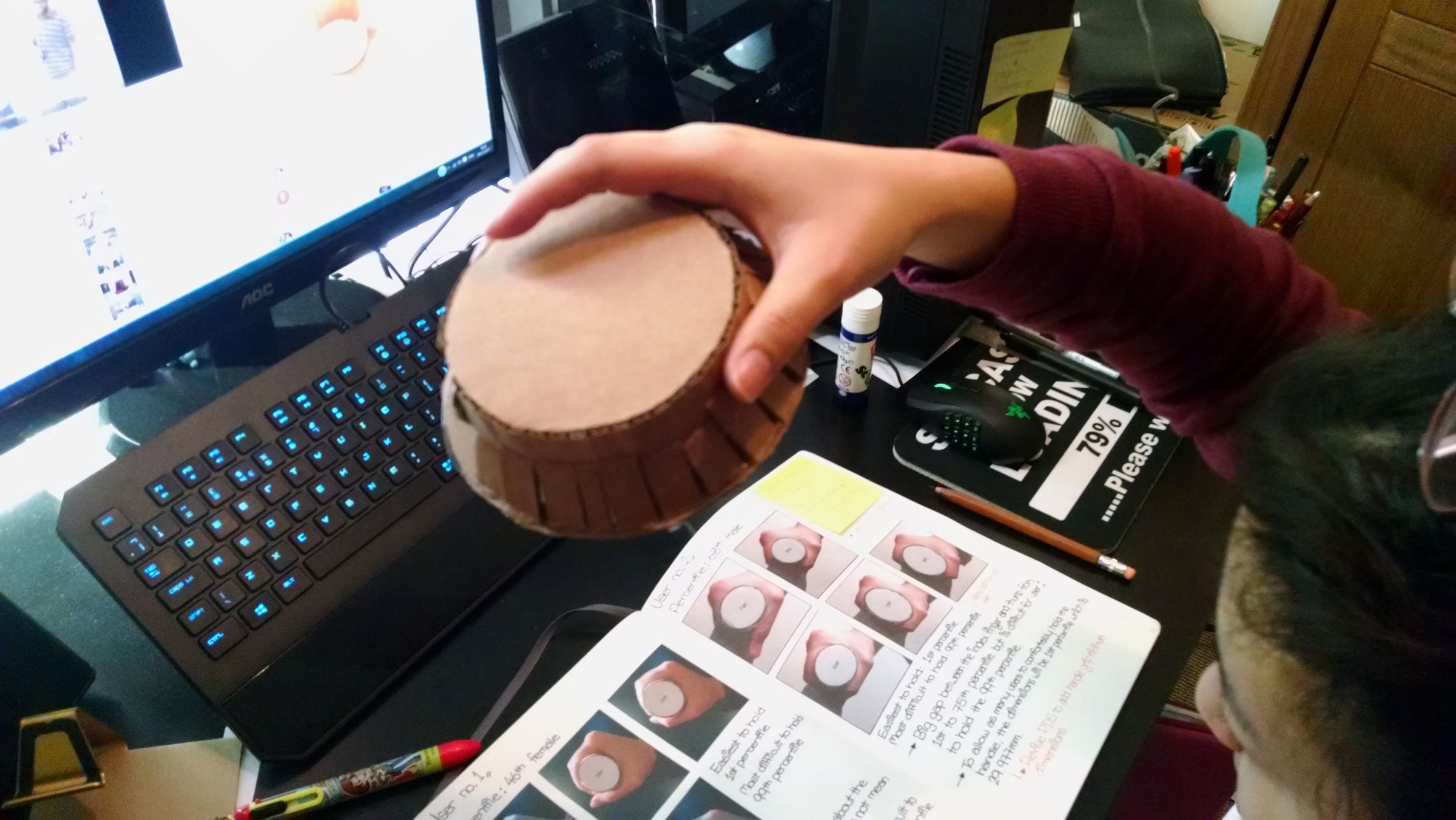 A hand holding a small turned wooden disc component next to a laptop, testing the fit of a dial or base part.