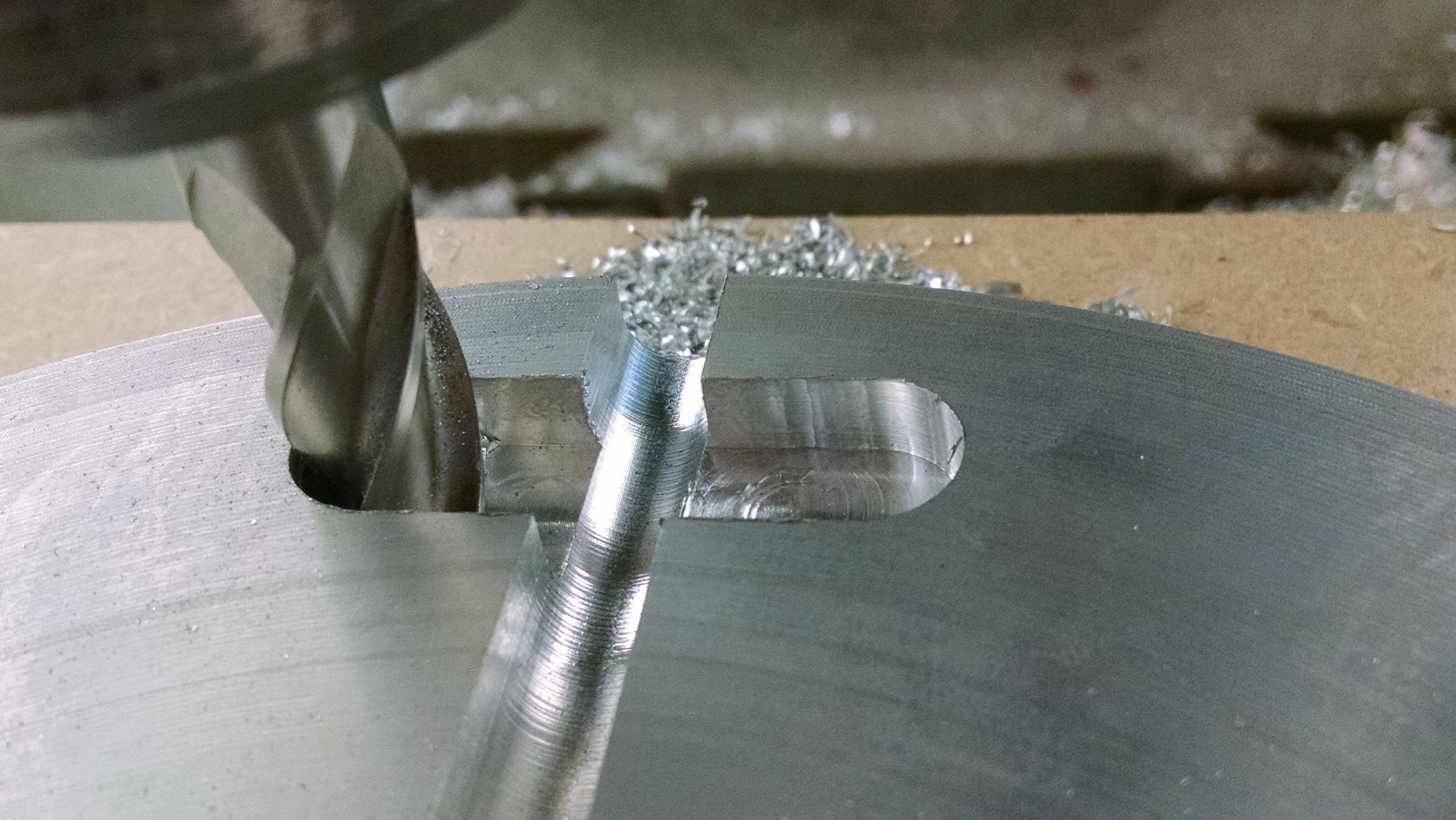 Close-up of a drill bit cutting into a block of material clamped to a workbench, with shavings scattered around.