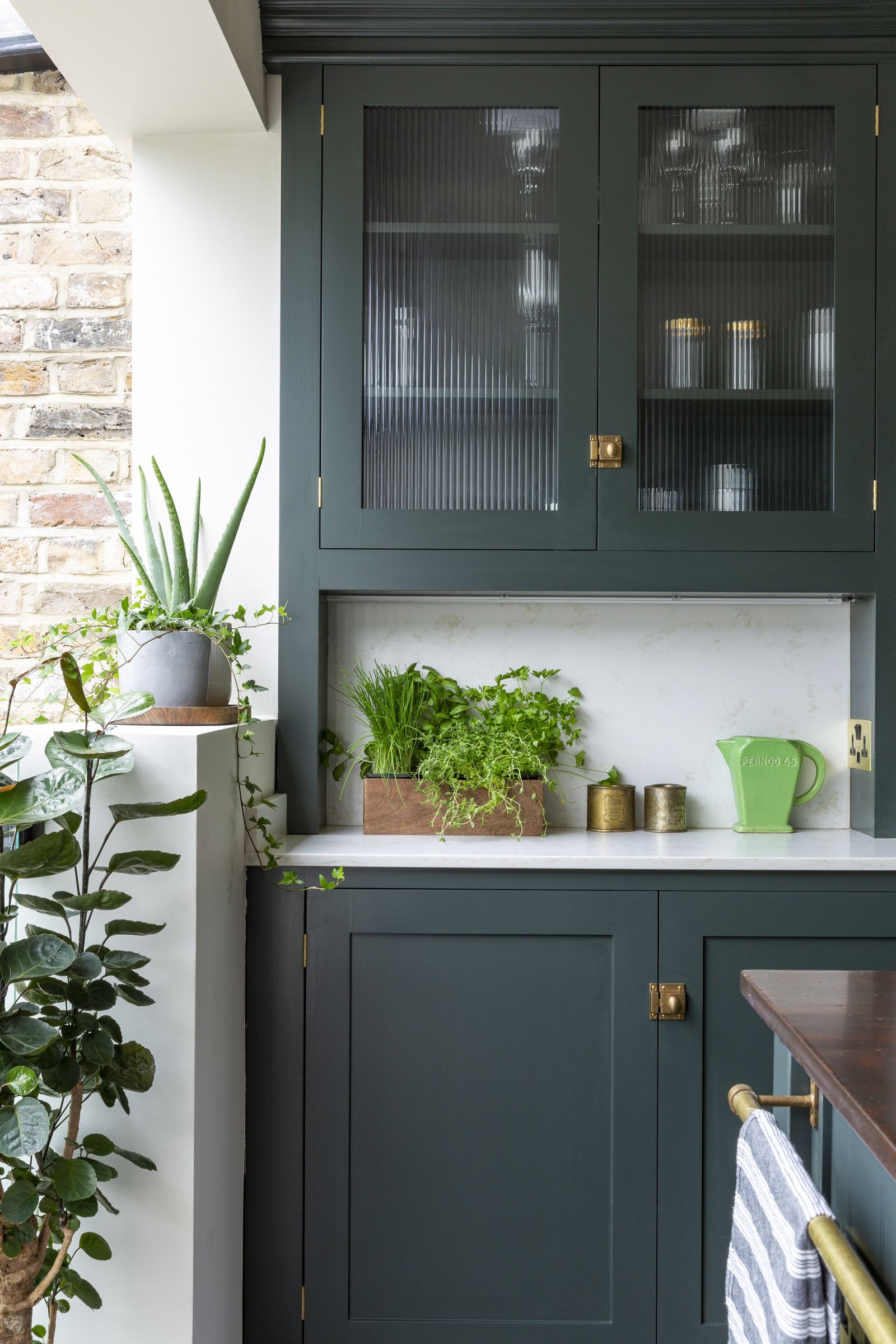 A tall dark green corner cabinet with glass-fronted doors, near a brick wall with potted plants on the counter below.