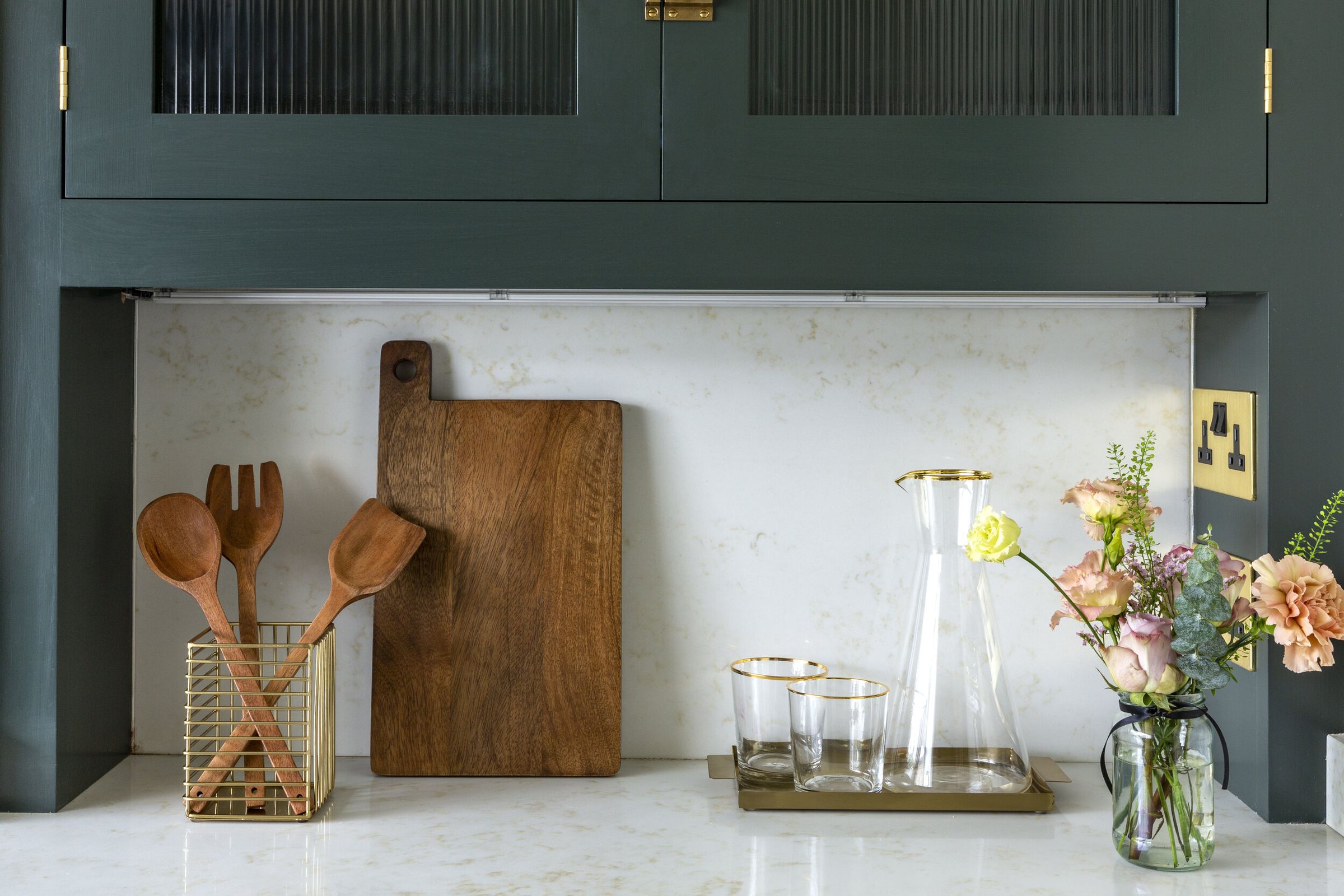 A styled countertop vignette showing wooden utensils in a caddy, a chopping board, and a small vase of flowers against a marble splashback.