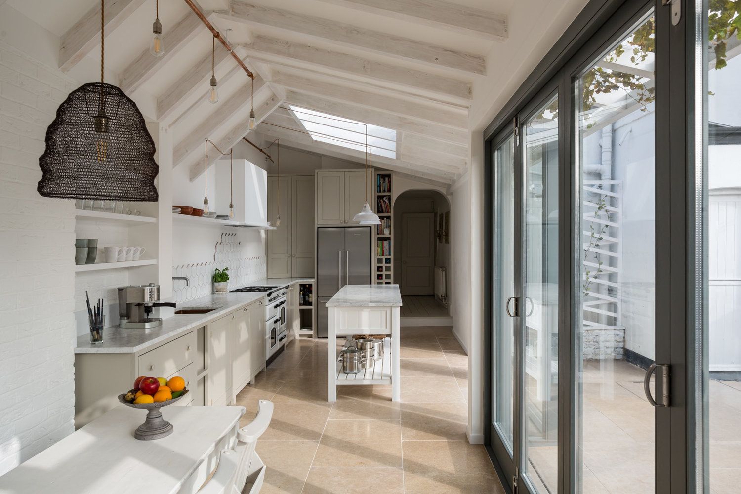 A long white kitchen extension with a vaulted glazed roof and a black woven pendant light, bifold doors open to the garden.