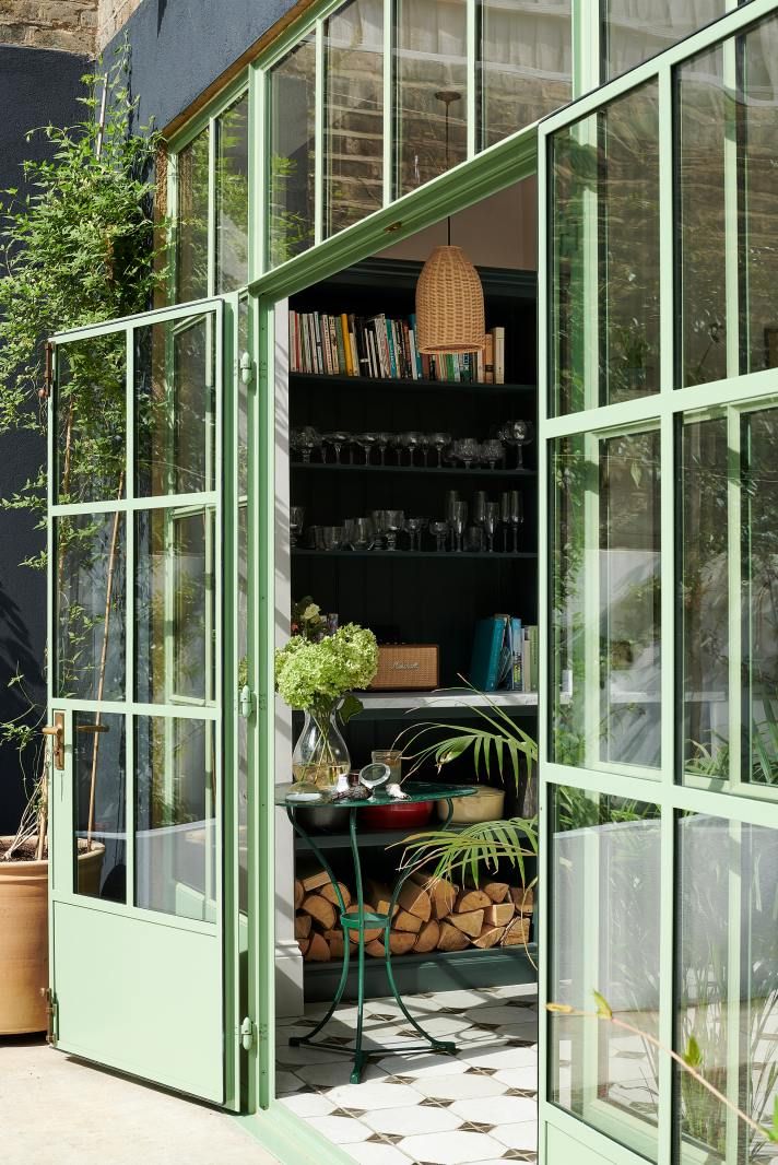 View through open green garden doors into a plant-filled outdoor seating area, with a black-and-white tiled floor inside.