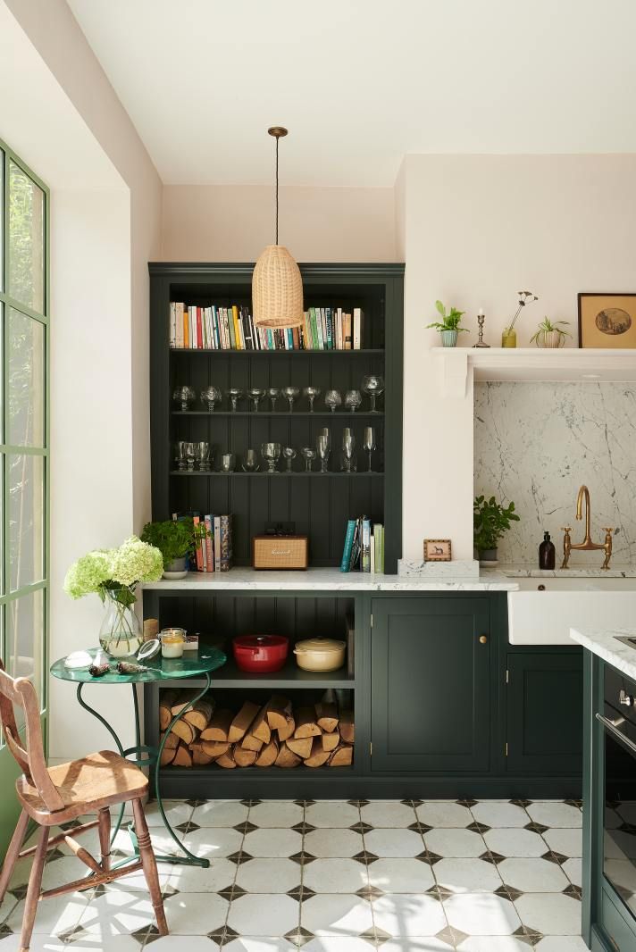 A dark green dresser unit with open shelving for glassware and books, log store below, blush pink walls.
