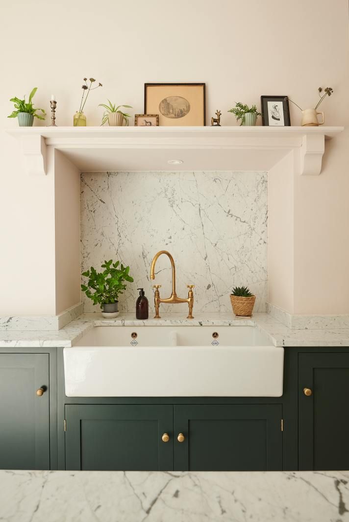A Belfast sink with brass tap set into dark green cabinetry, marble splashback, and a mantel shelf styled with plants and framed pictures above.