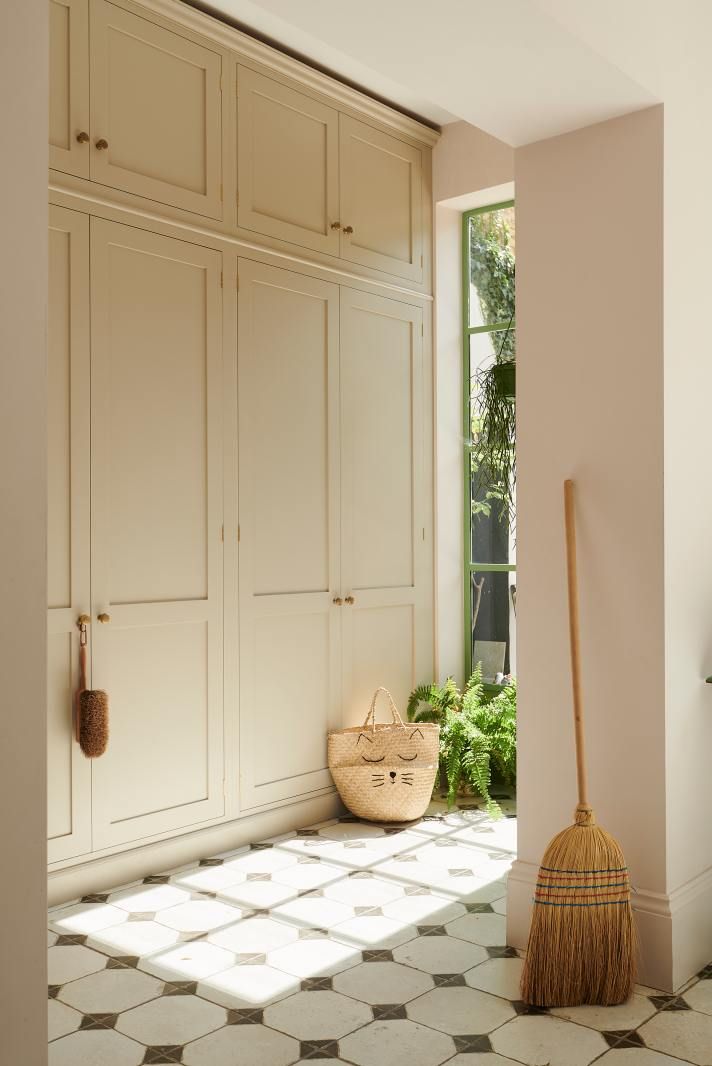 Tall built-in cabinetry in soft beige, with a broom leaning against it and sunlight falling across the monochrome tiled floor.